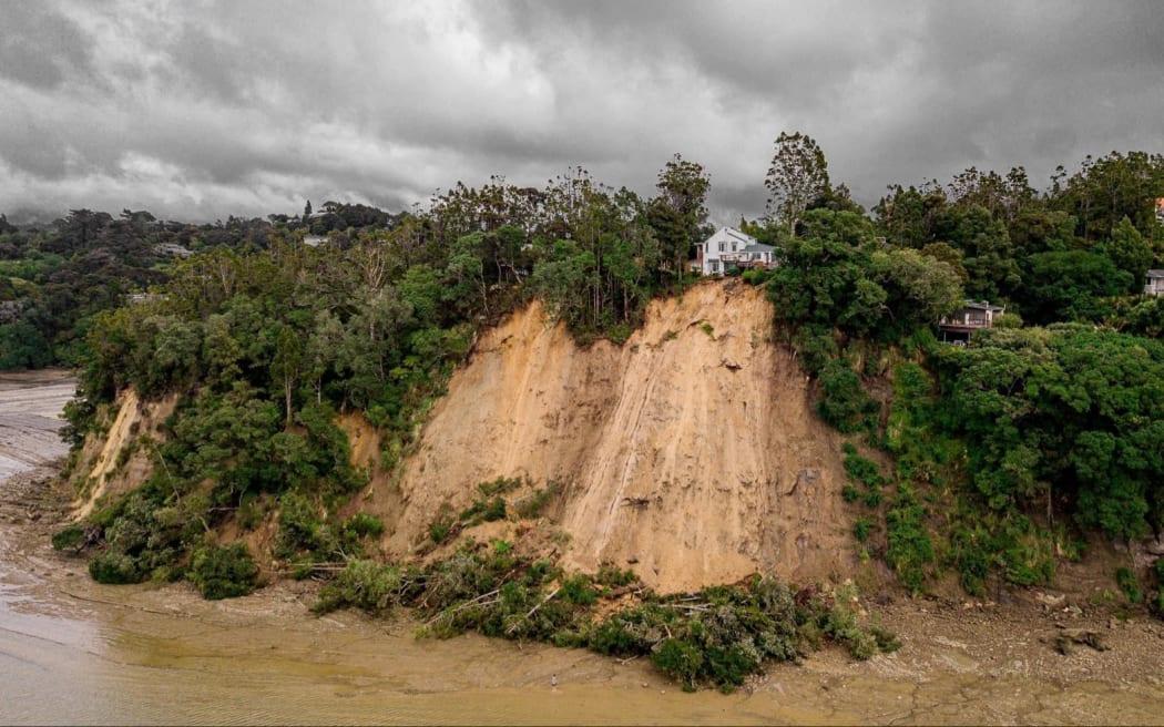 A wide view of a large landslide showing trees having slipped down a steep cliff and a house perched precariously on top.
