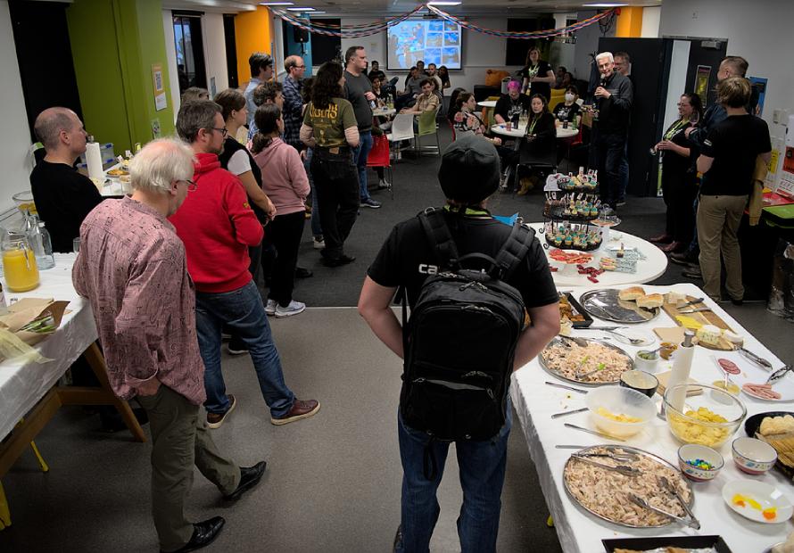 A crowd of people standing and sitting in a large room at the end of the day. Someone is talking on a microphone and there are tables of food in the foreground.