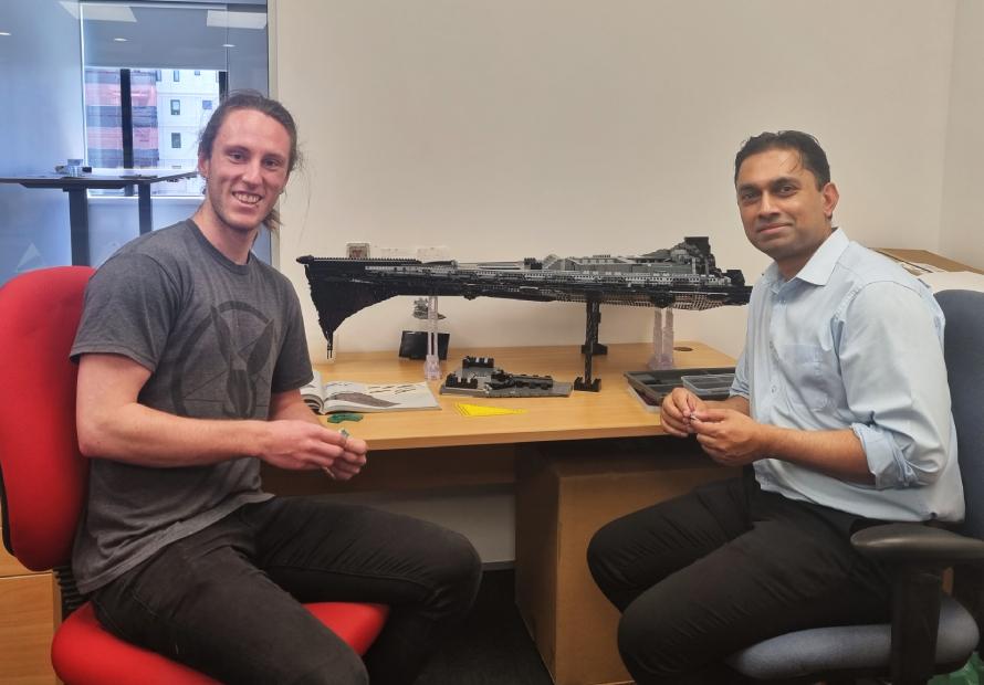 Liam Sharpe and Sanjay D'Souza are sitting on chairs in a brightly lit office with a large, unfinished Star Wars Lego set on the table behind them.”
