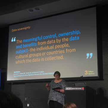 Kathy Reid pictured while presenting at Everything Open 2025. The screen behind her displays a definition of data sovereignty “the meaningful, control, ownership, and benefits from data and by the data subject – the individual people, cultural groups or countries from which the data is collected.”