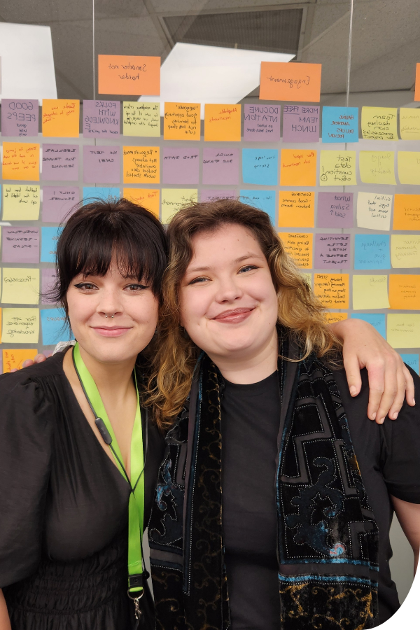 Fyfa's arm wraps around Maria's shoulders as they smile in front of a wall of post it notes