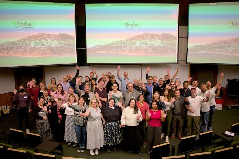 A large group gathers infront of projectors. They smile and raise their arms up.