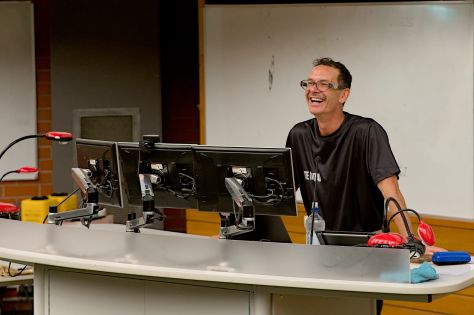 Peter-Lucas Jones stands behind a desk, resting his hands on it as he speaks. He is smiling and is wearing glasses.