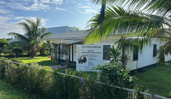 Photo of the Kolovai Community Public Library. In front of the library is a welcome sign display ‘Ko e Fale Laukonga’ with information about what’s available.  
