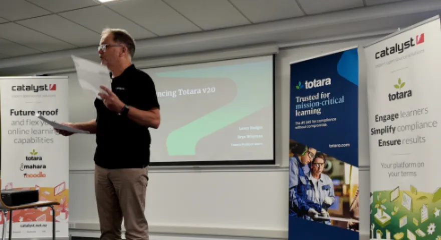 Shayne Priddle stands infront of a presentation with a paper in hand. Behind him are Catalyst and Tōtara banners