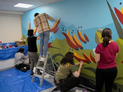 Several people including one person on a ladder are painting a mural on a wall in a meeting room with bright blue green red and yellow colours