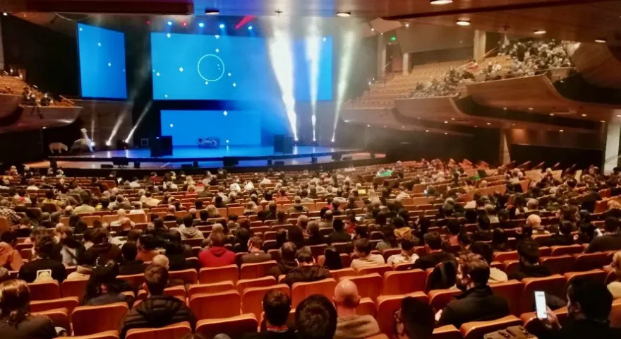 A huge event room with hundreds of people seated looking at a blue lit stage