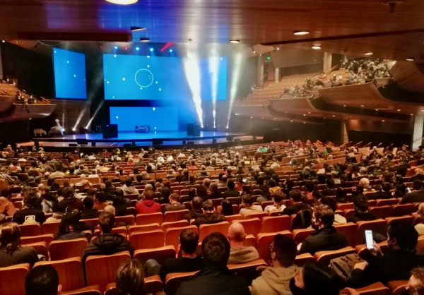 A huge event room with hundreds of people seated looking at a blue lit stage
