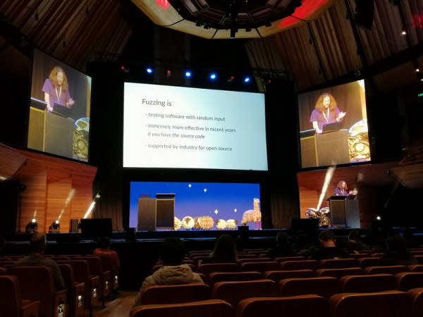Douglas Bagnall is presenting a talk on stage at Kawaiicon with large screens behind him.