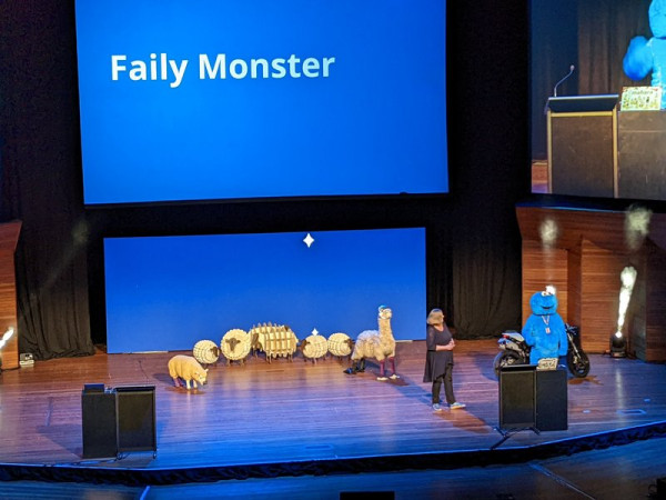 Kristina Hoeppner is on stage with someone in a large fuzzy blue creature costume and a line up of (not real) farm animals.