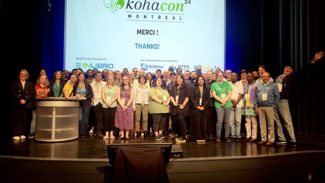 KohaCon24 attendees at the end of Day 2 taking a group photo on the conference stage.