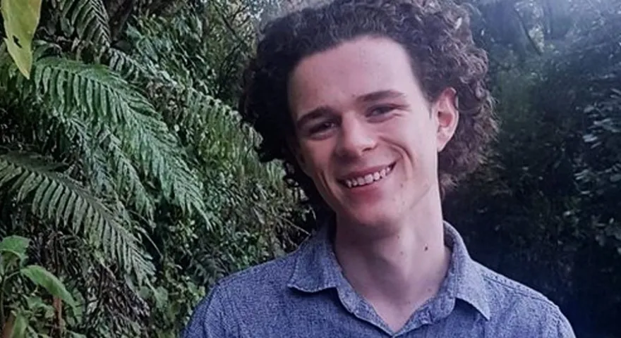 Daniel Southward Ellis is standing in front of some ferns wearing a blue button up shirt and smiling at the camera