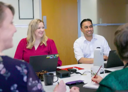 A group of people smiling, the camera focuses on a woman in a pink smiling sat next to a man in a white shirt