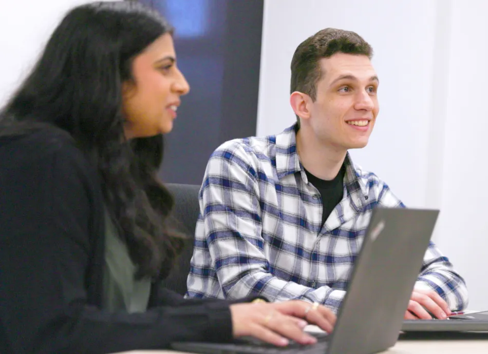 A woman typing on a laptop sat next to a man in a check shirt. 