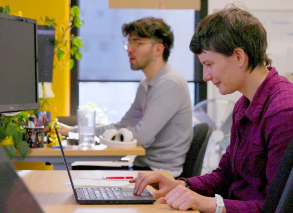 Two people with short hair working at a desk on their laptops and computer screens