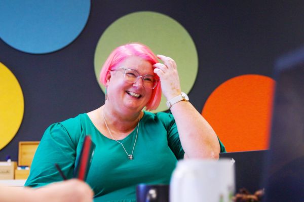 Katherine Ryan has bright pink hair that she brushes off her face as she smiles. She wears glasses and a teal top. Behind her, are a wall of bright polka dots.