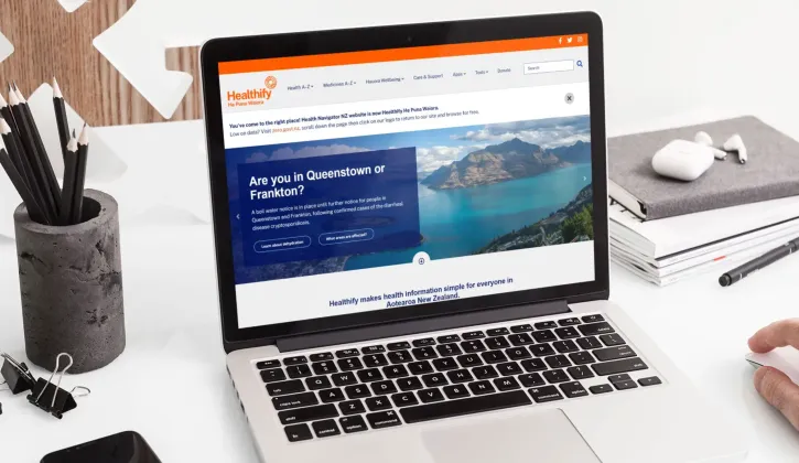 A laptop sits on a desk with stationery items around it displaying a screenshot of the Healthify website which asks are you in Queenstown or Frankton