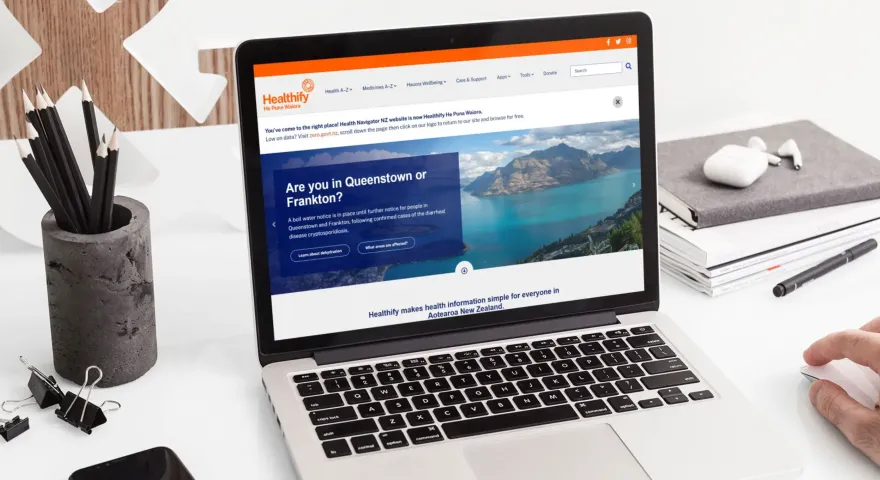 A laptop sits on a desk with stationery items around it displaying a screenshot of the Healthify website which asks are you in Queenstown or Frankton