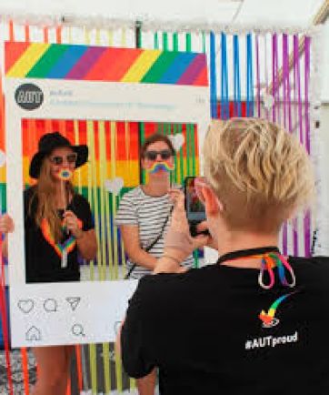 A person with short blonde hair taking a photo of two other people who are stood infron of a rainbow themed backdrop