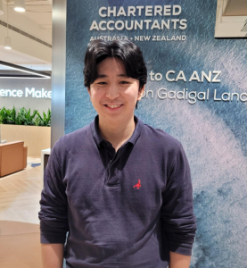 Smiling headshot of Ed. He wears a blue polo shirt and stands infront of a wall with the CA ANZ sign
