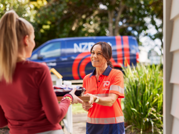 A NZ Post courier, smiles as she hands over two parcels to a woman. In the background is a NZ Post van