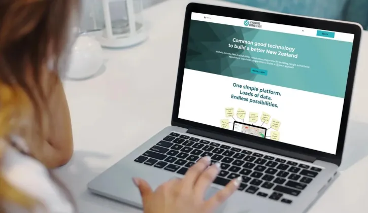 A person with long hair sitting at a desk looking at a laptop on the National Forward Works Viewer website homepage that showcases Common good technology to build a better New Zealand and the op