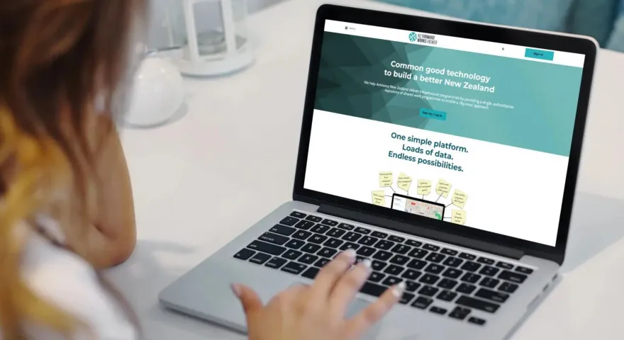A person with long hair sitting at a desk looking at a laptop on the National Forward Works Viewer website homepage that showcases Common good technology to build a better New Zealand and the op