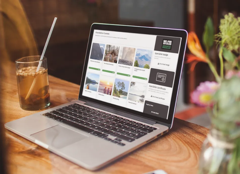 An open laptop displays the foundation courses for travel sellers in Tōtara. On the table next to it is an iced coffee and a vase of flowers.