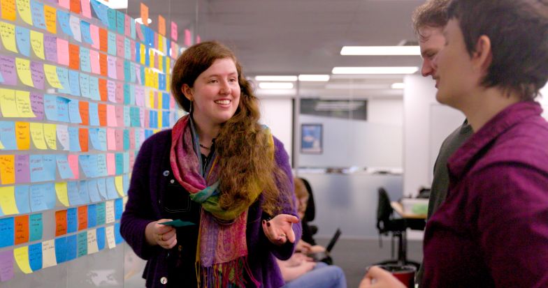 A woman with long ginger hair wearing a purple cardigan smiling stood infront of a wall of Post-its