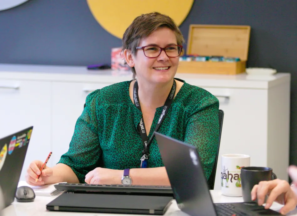 A woman with short hair in a green shirt sat at an office table smiling