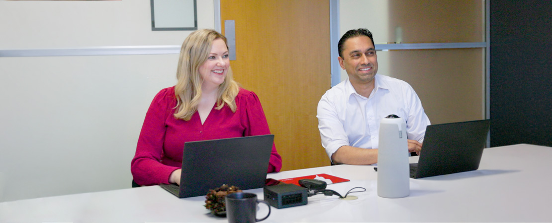 Homepage Hero Two office workers sit around a desk with laptops smiling