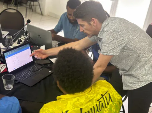 Tim Beale leans over a table and points at an open laptop as he teaches someone how to use RiskScape.