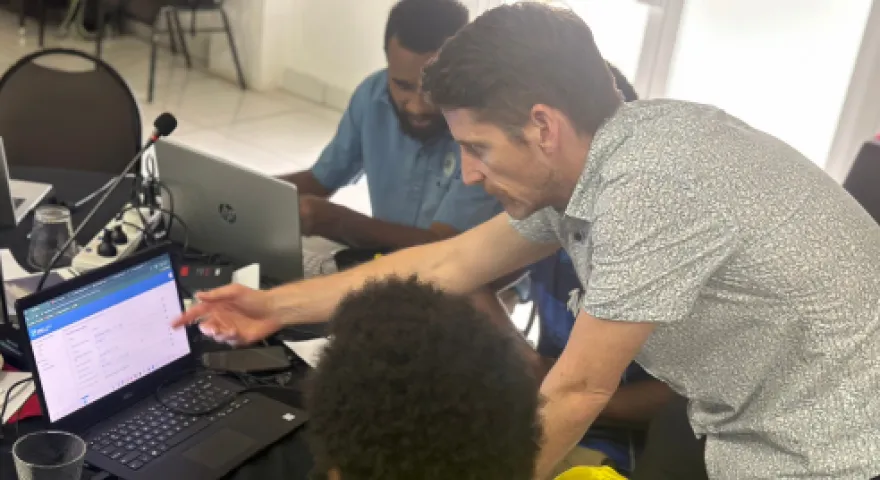 Tim Beale leans over a table and points at an open laptop as he teaches someone how to use RiskScape.