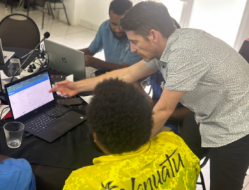 Tim Beale leans over a table and points at an open laptop as he teaches someone how to use RiskScape.